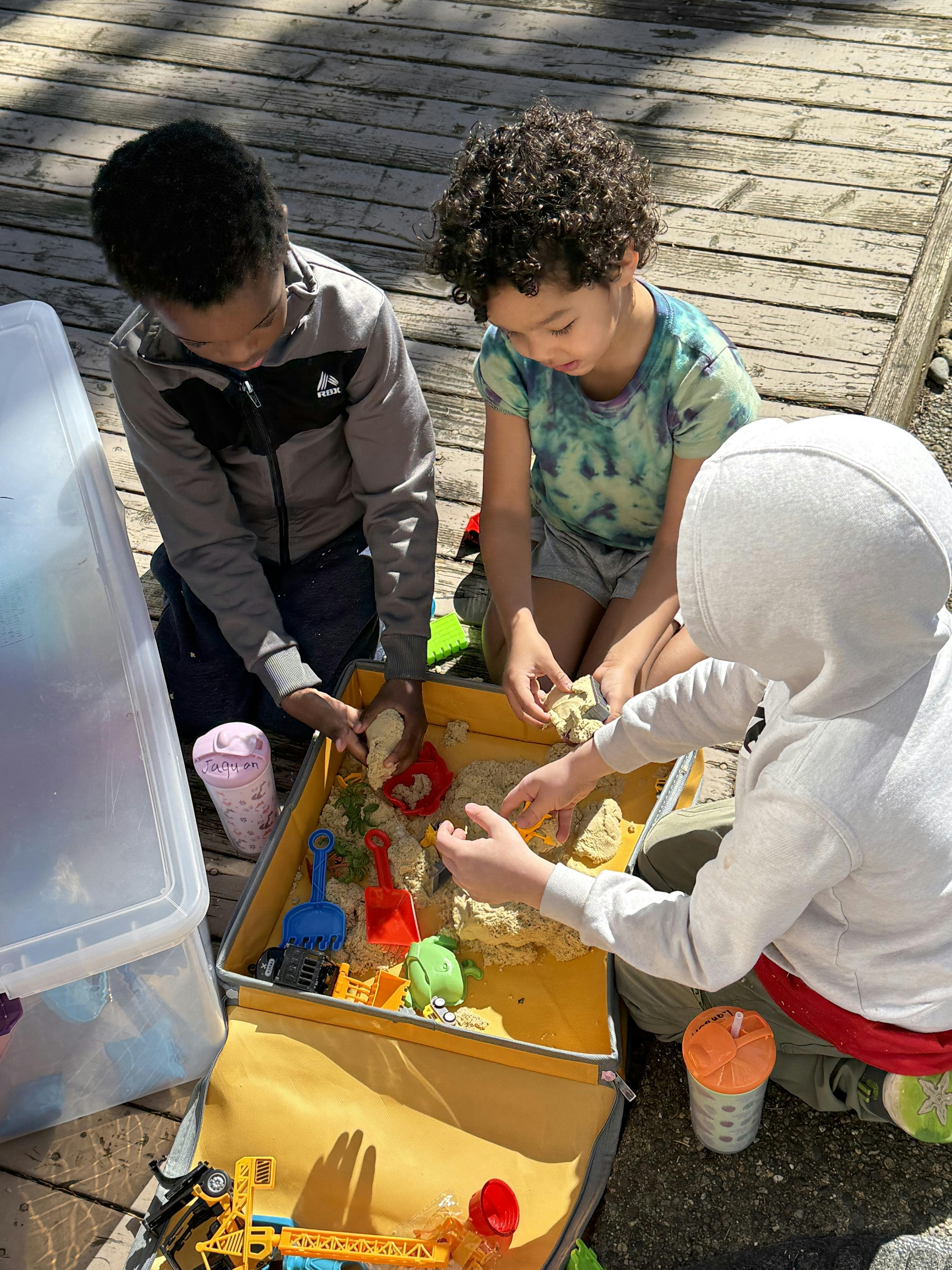 Three children playing with kinetic sand and toys in a yellow container