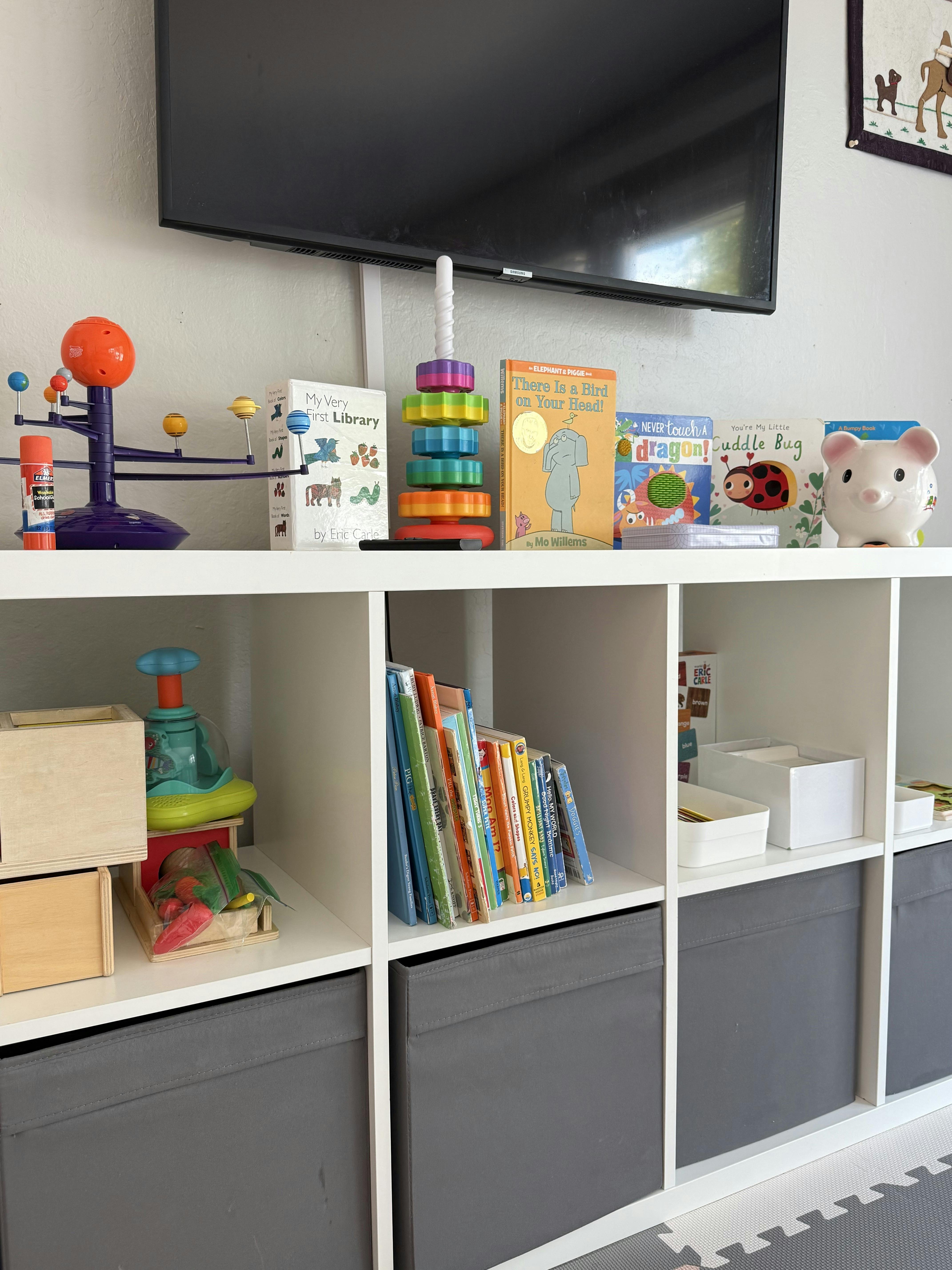 White toy storage shelf with children's books and toys beneath a wall TV