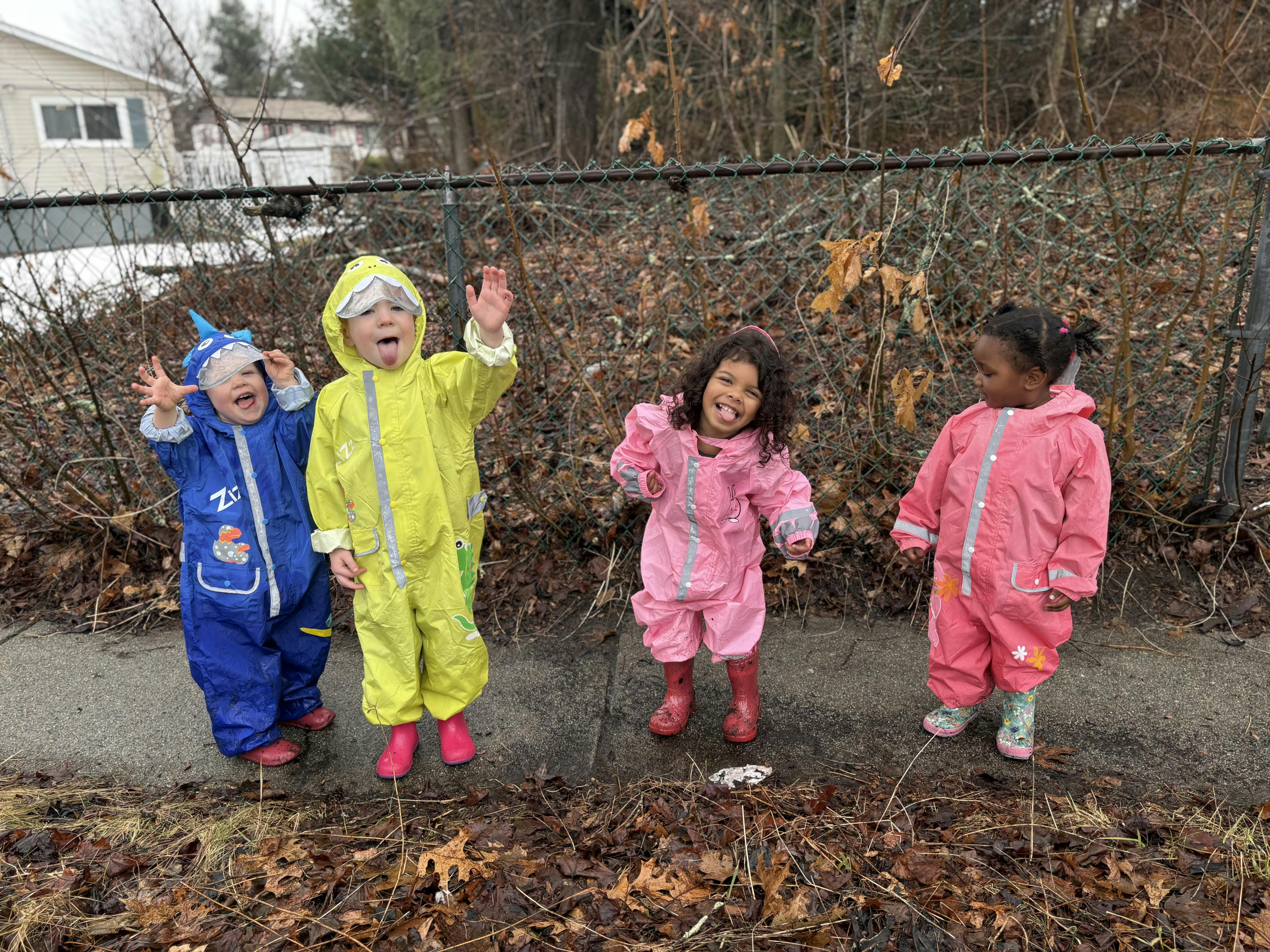 Four children in colorful rain suits standing next to a fence