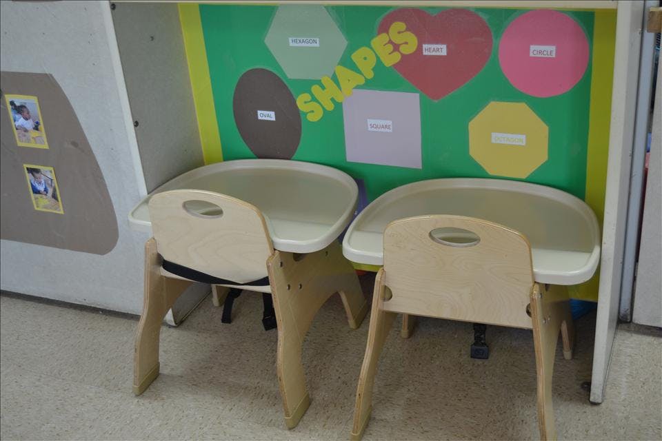 Two child-sized chairs with trays facing a colorful shapes learning board