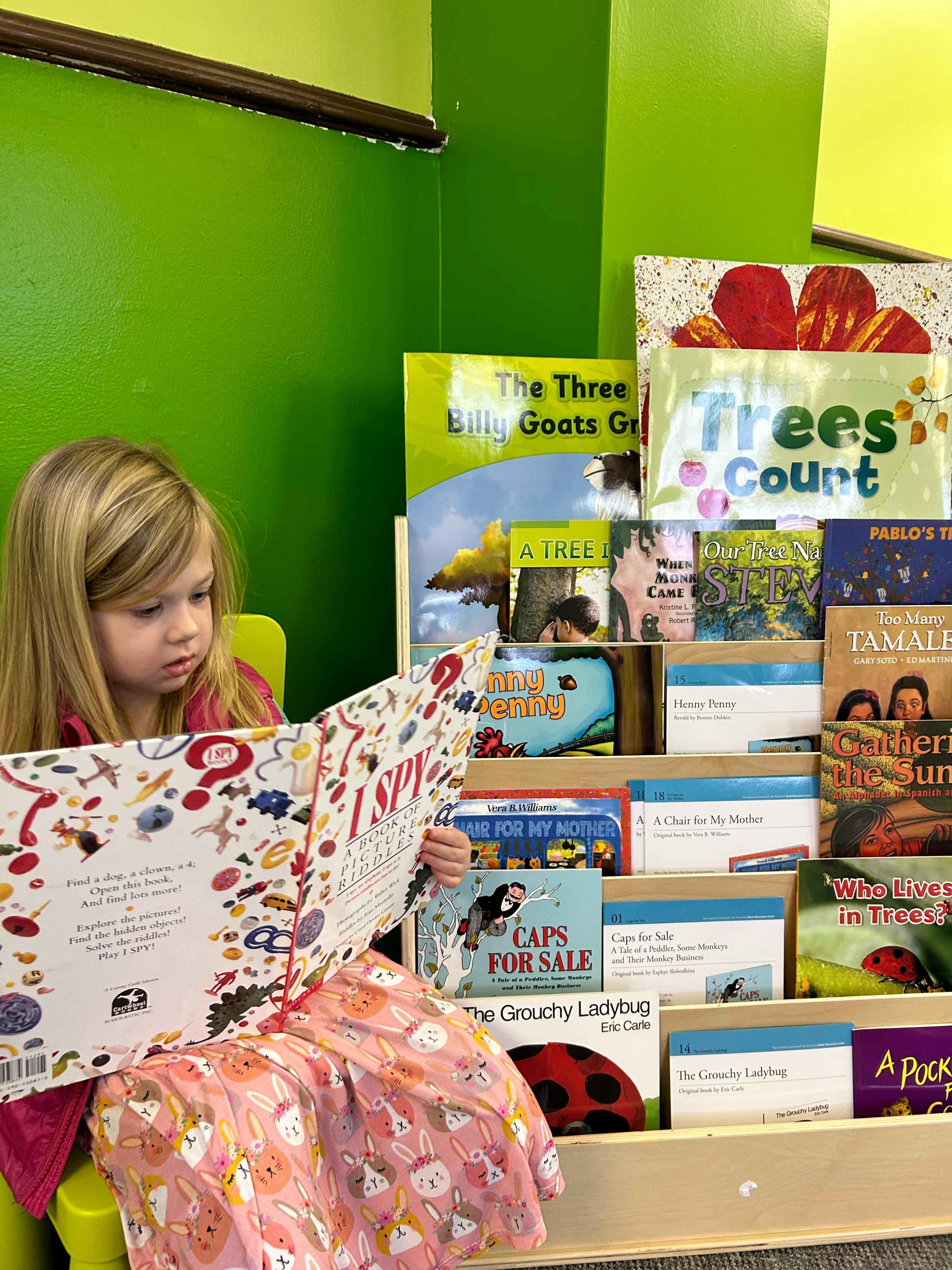 Child reading a large book in front of a bookshelf filled with children's books