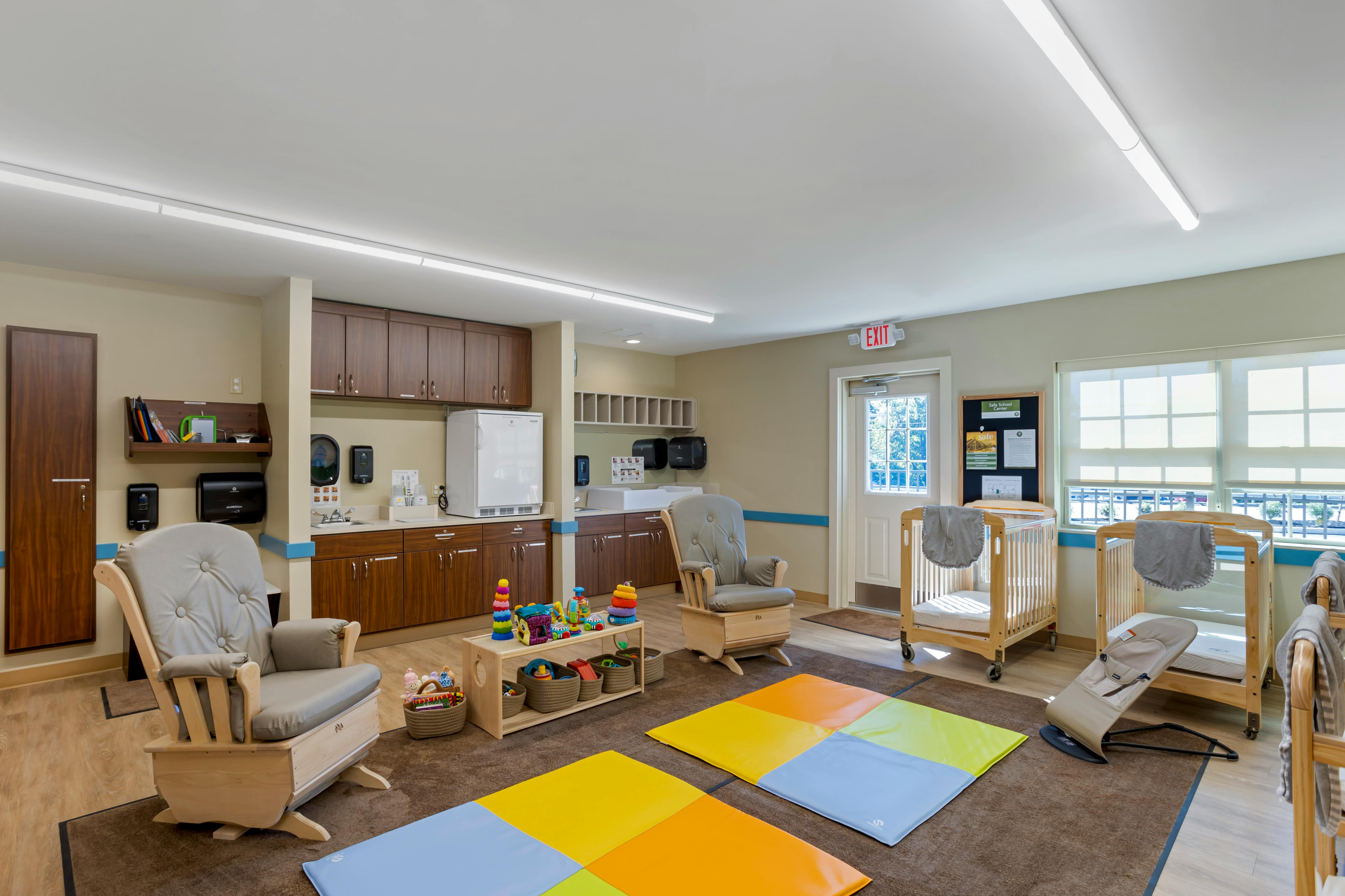 A childcare room with rocking chairs, cribs, changing stations, and colorful floor mats