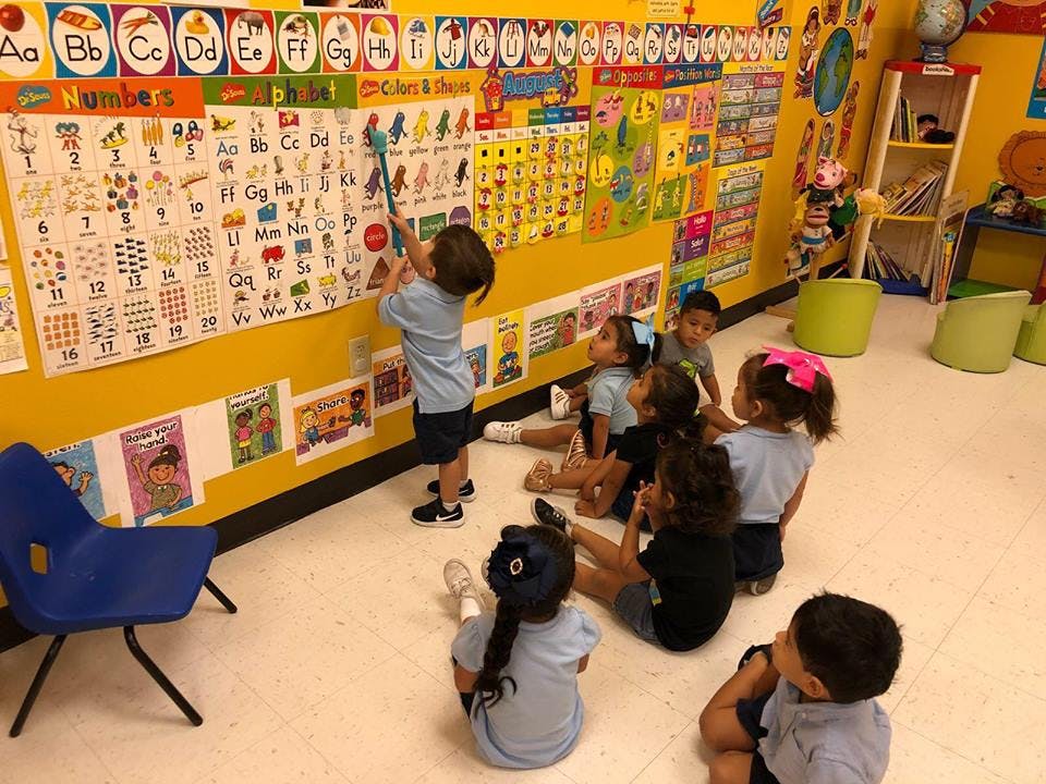 Child pointing at an alphabet chart in a classroom with other children watching