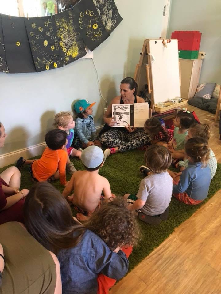 Person reading a book to a group of children sitting on a green rug