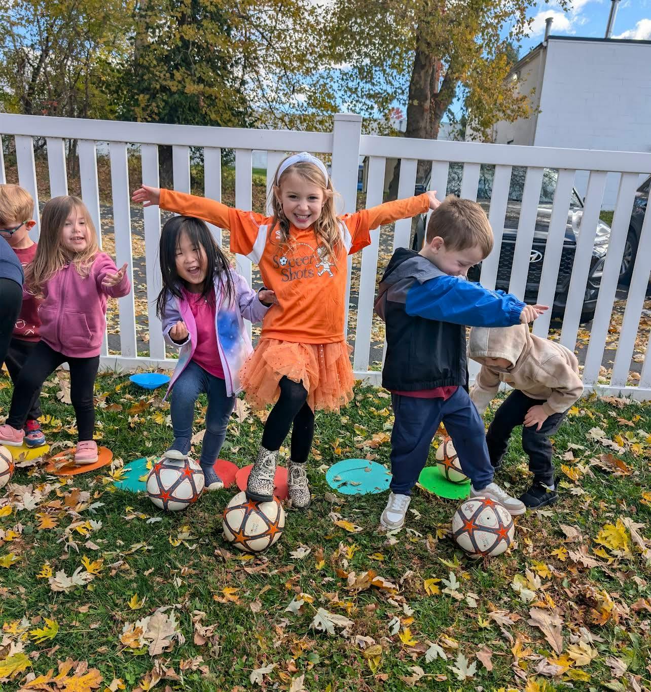 Group of children standing with soccer balls on a grassy area with fallen leaves