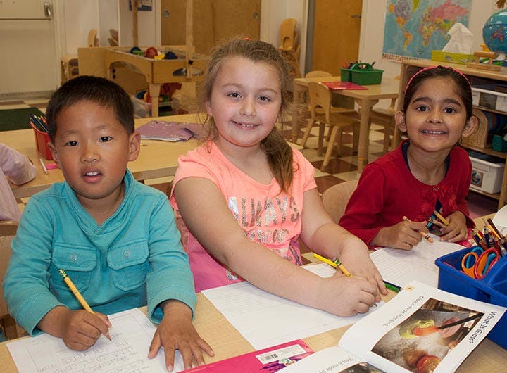 Three children drawing and writing at a table in a classroom