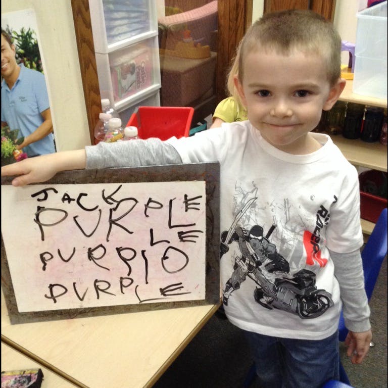 Child standing next to a sign with the word "PURPLE" written on it