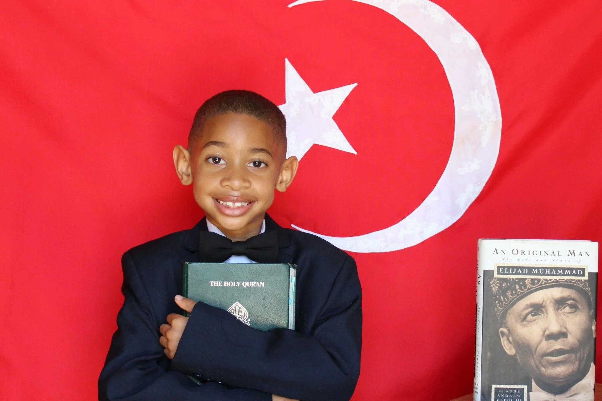Child in a suit holding a book titled "The Holy Qur'an" with a red flag behind