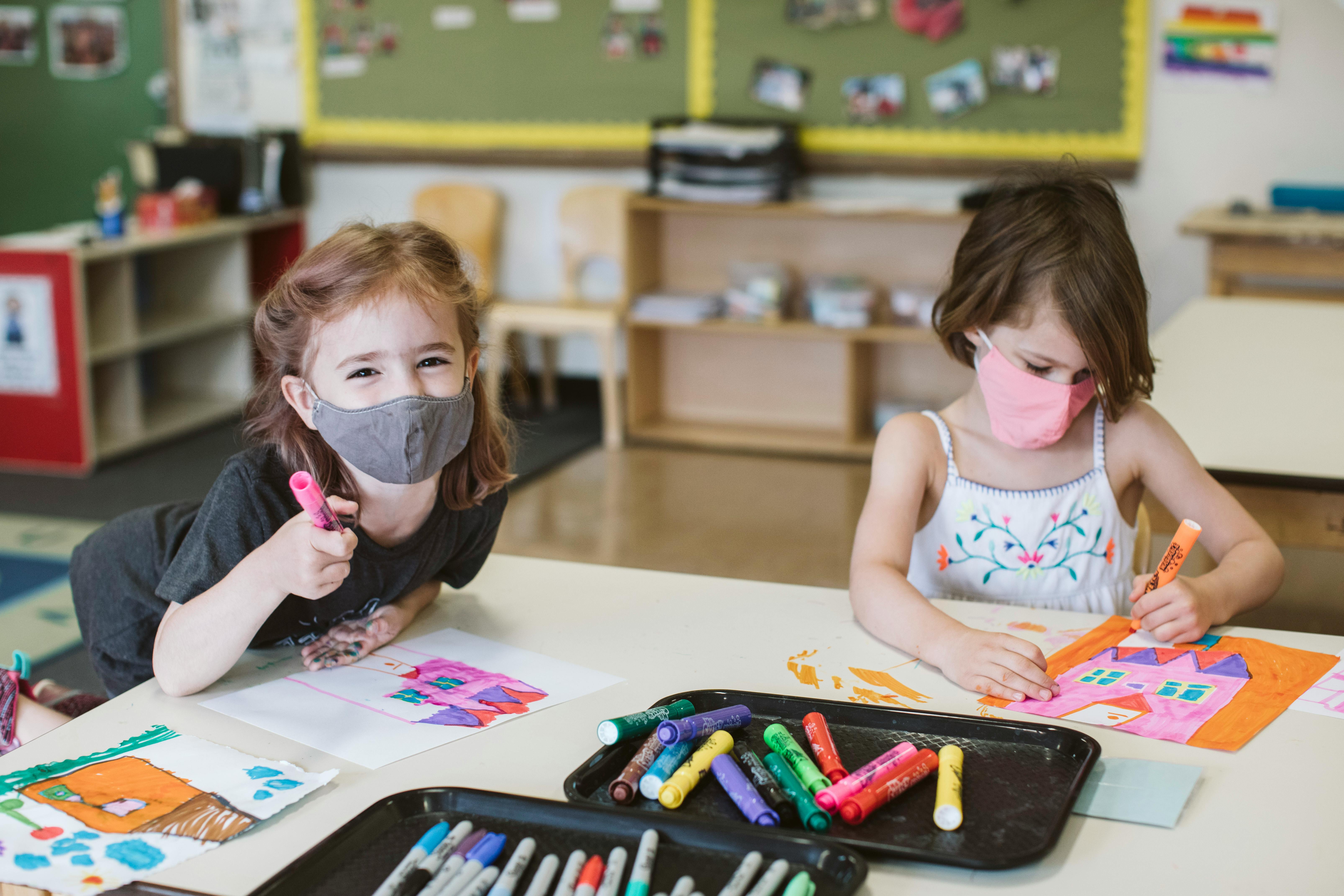 Two children wearing masks drawing at a table in a classroom