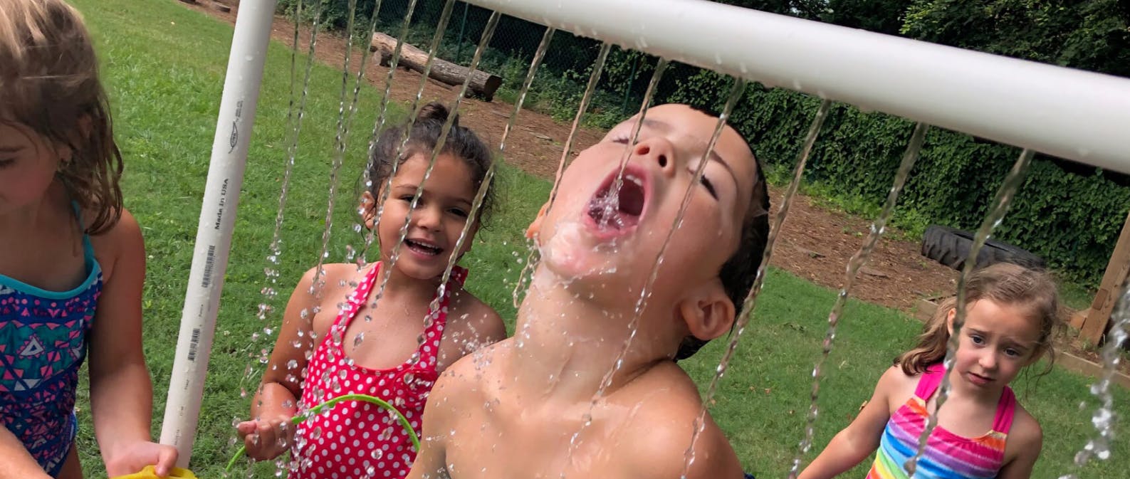 Child with mouth open under falling water, other children nearby