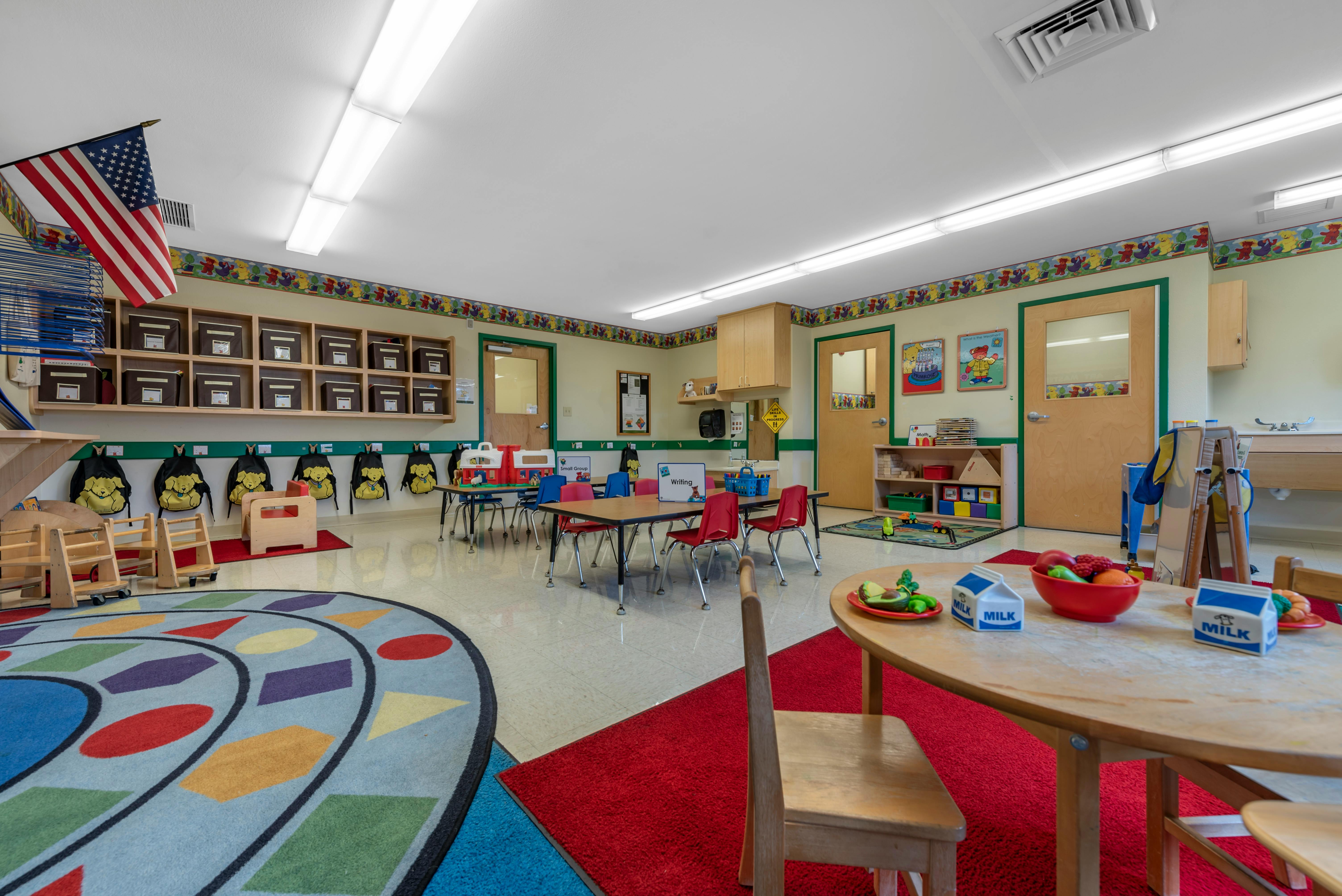 A colorful classroom with small tables and chairs, toys, and an American flag