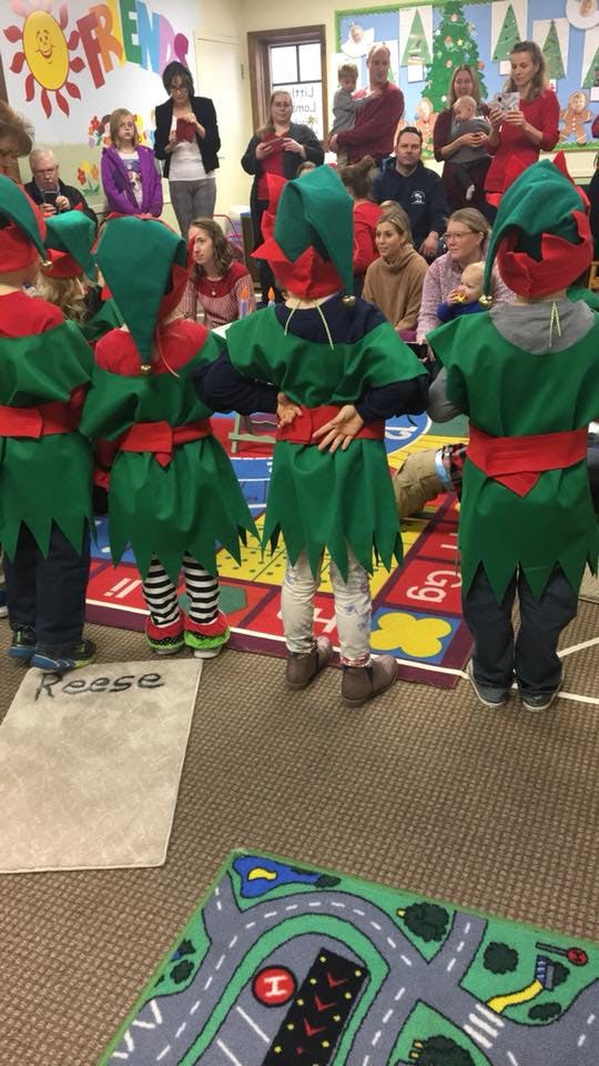 Children in elf costumes facing away from the camera, with adults in the background