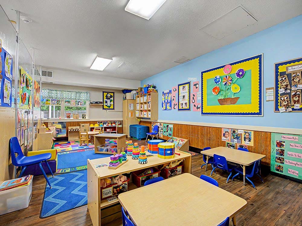 Colorful preschool classroom with tables, chairs, rugs, and artwork on the walls