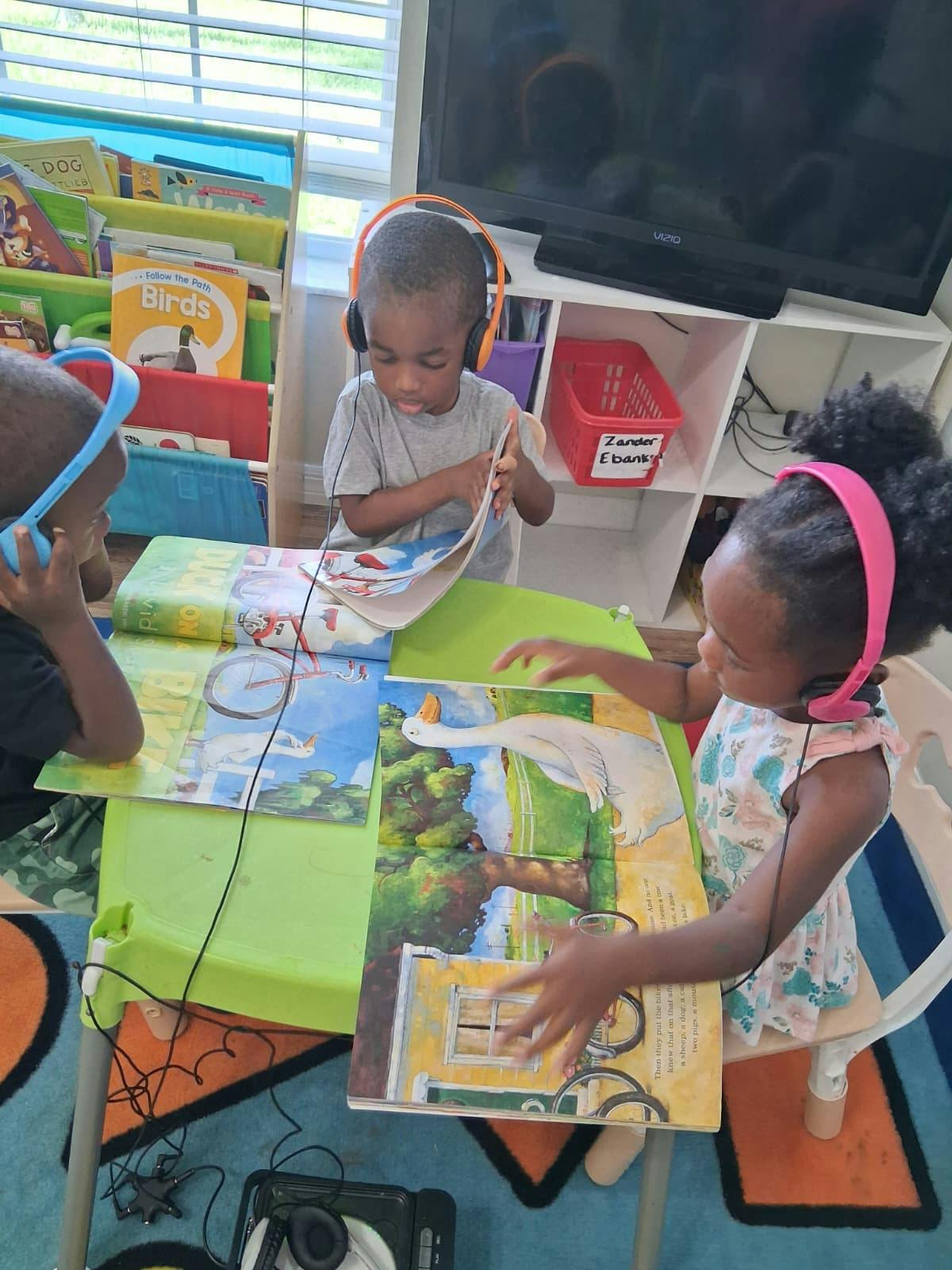 Three children wearing headphones reading books at a table