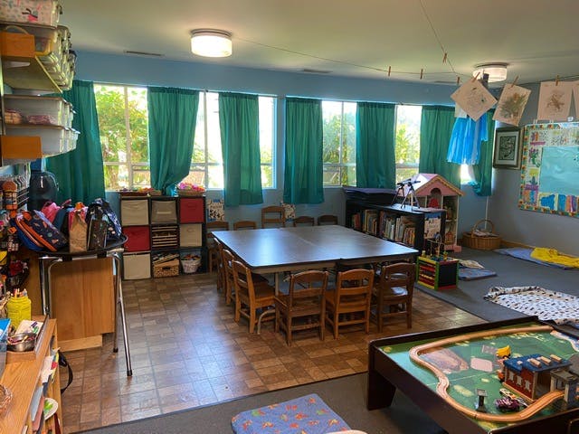 Classroom with a large table and chairs, shelves, and windows with green curtains