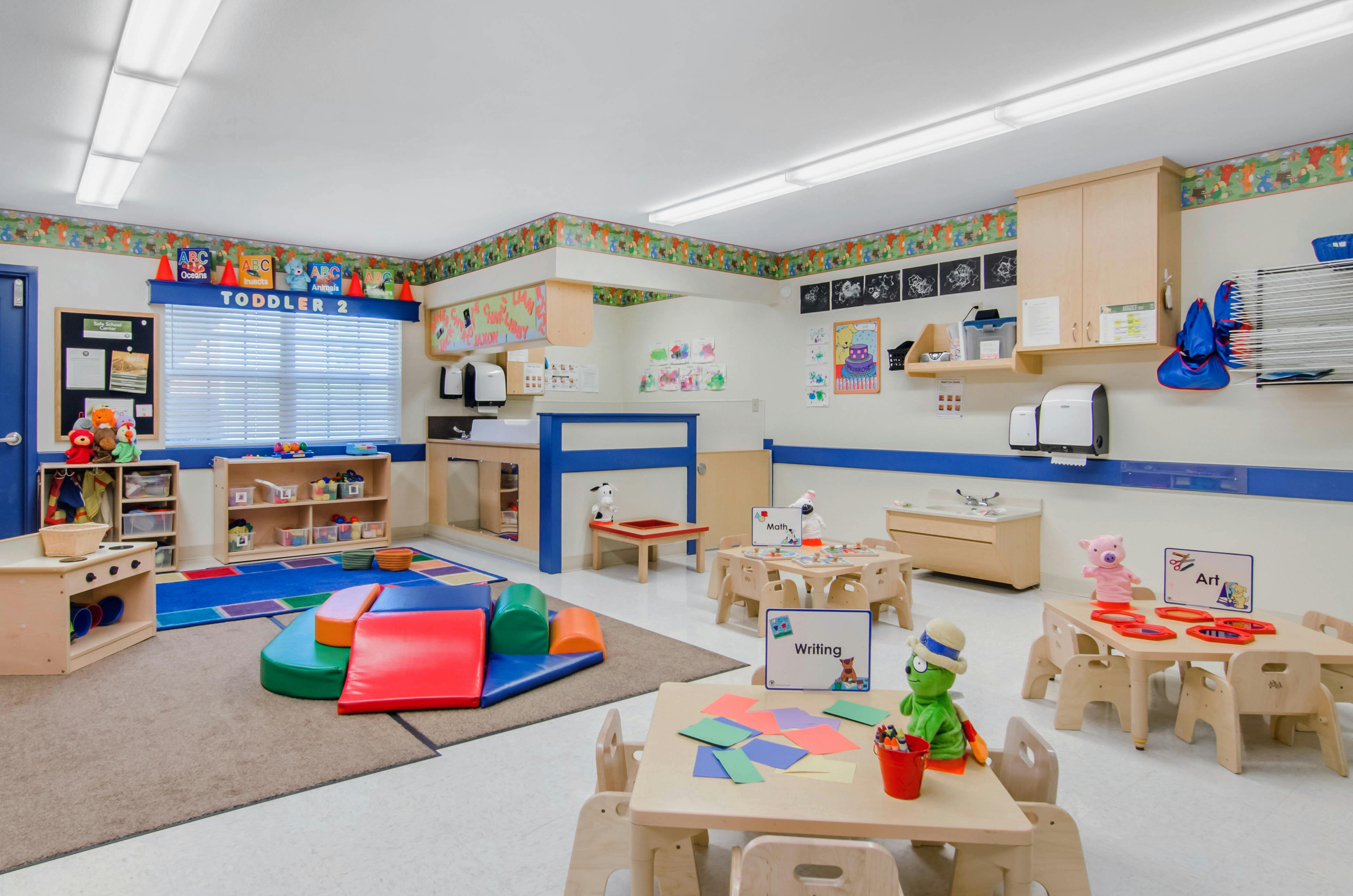 A colorful toddler classroom with small tables, chairs, and play mats