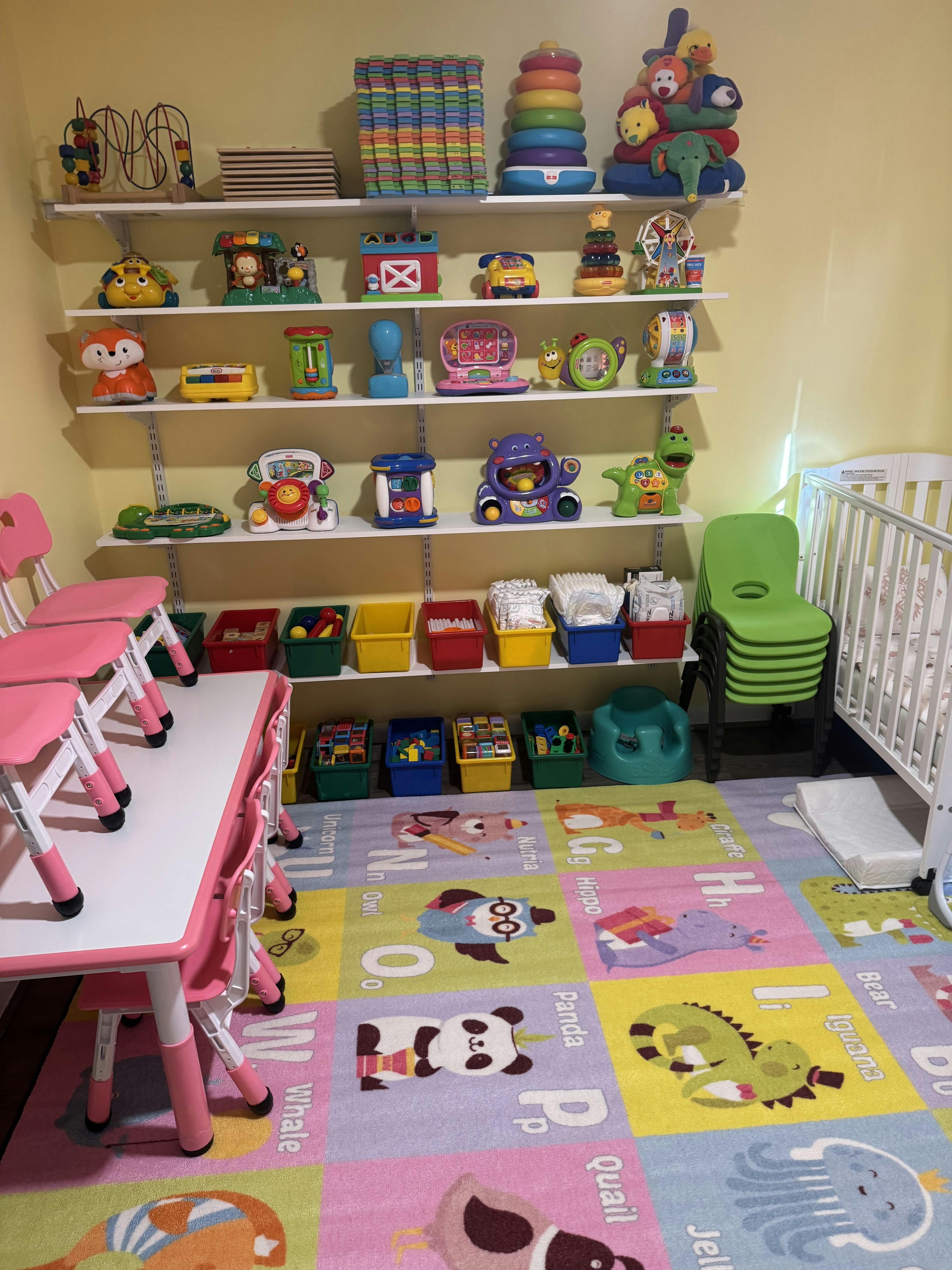 Childcare room with shelves of toys, small pink table and chairs, crib, and alphabet rug