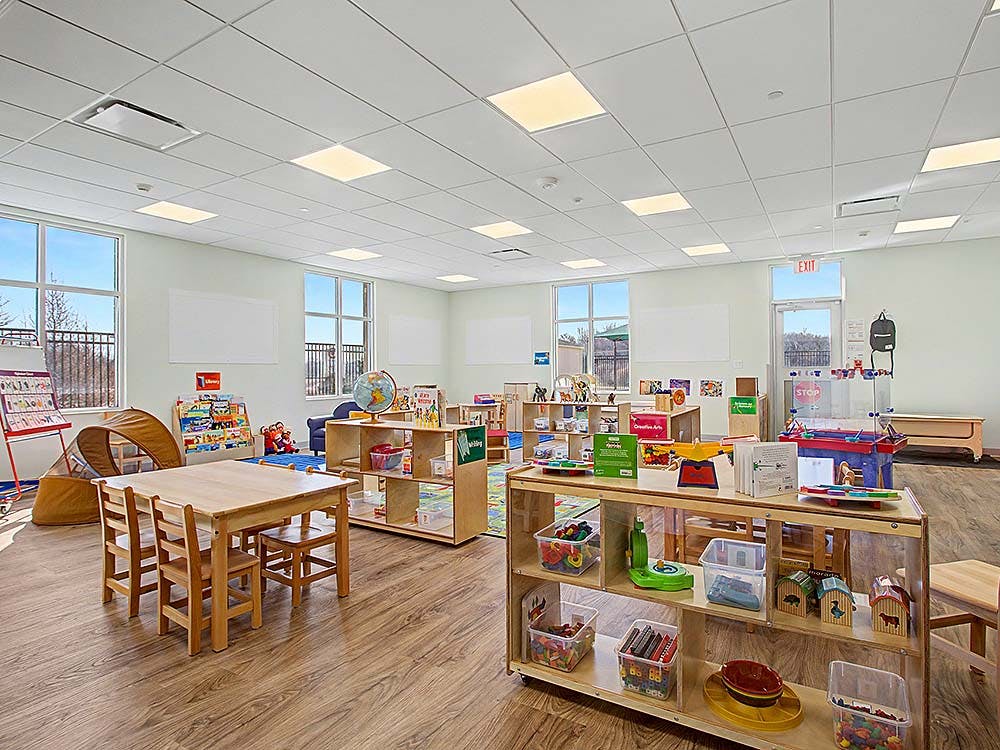 Empty children's classroom with small wooden tables, chairs, and shelves of toys
