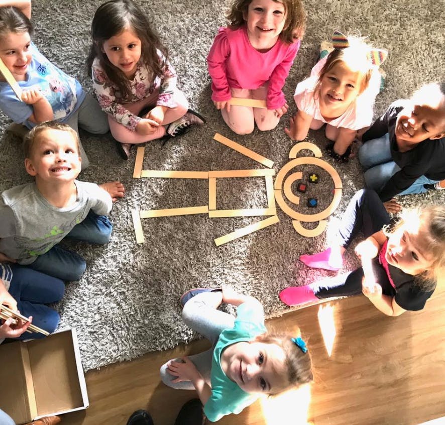 Children sitting on a rug around wooden blocks arranged to form a shape