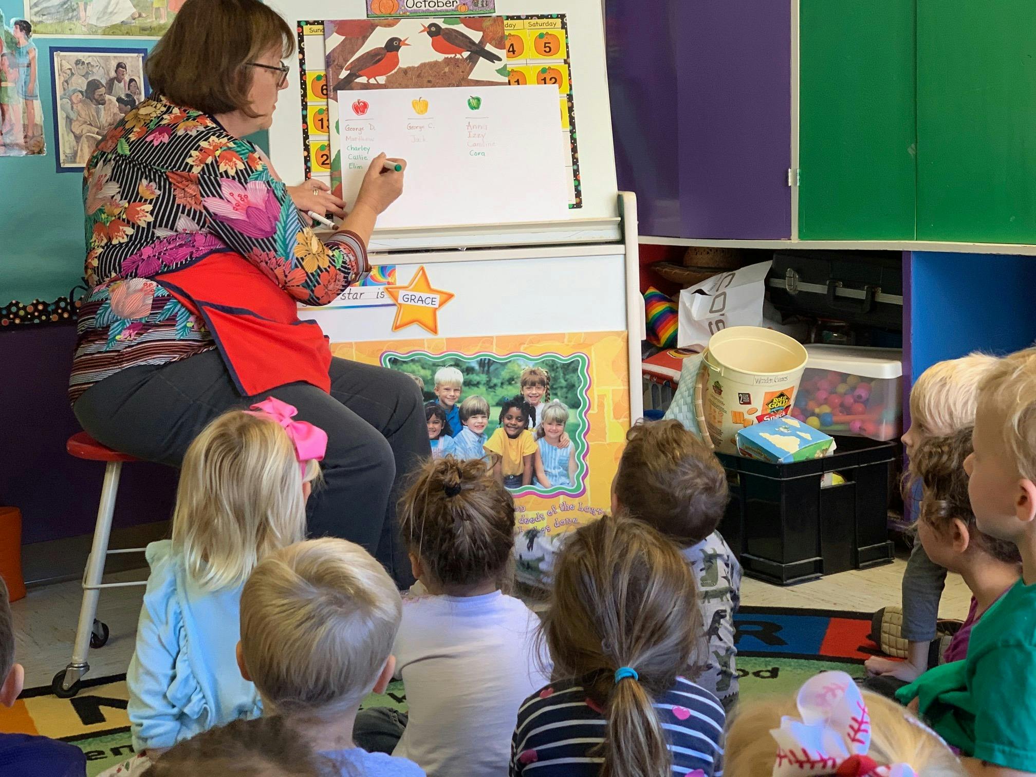 Person writing on a whiteboard in front of children sitting on the floor