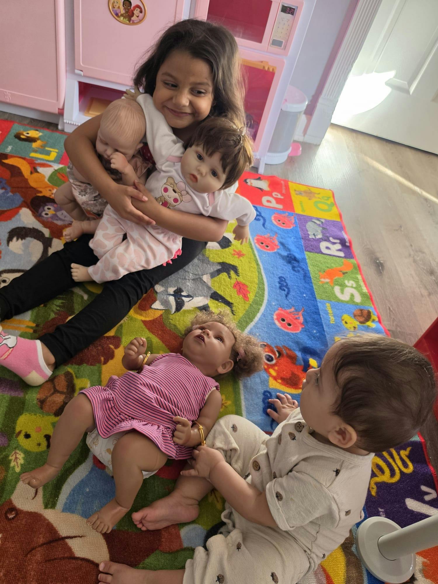 Child holding two dolls while another child plays with a doll on a rug