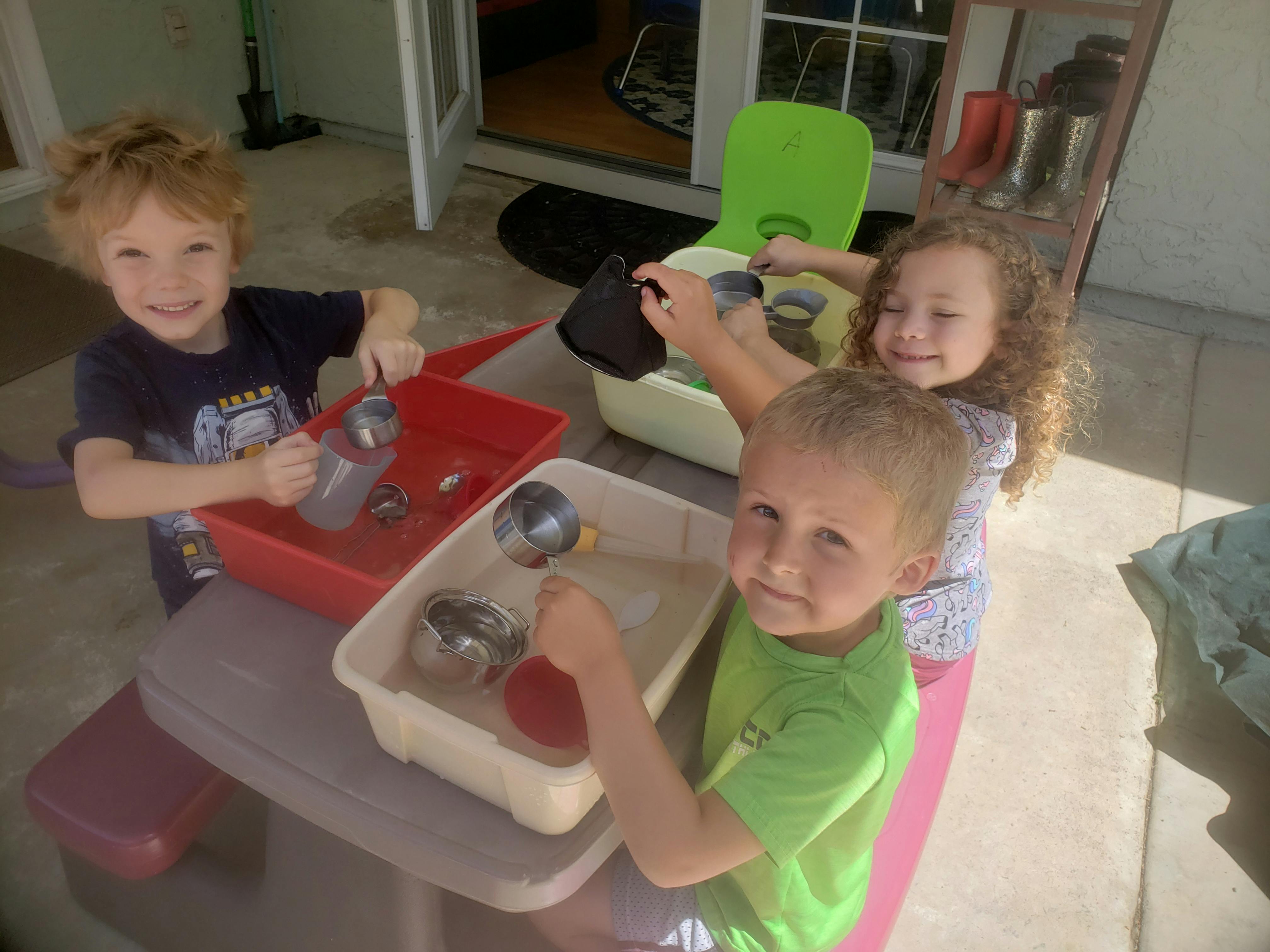 Three children playing with water and containers at a table with containers