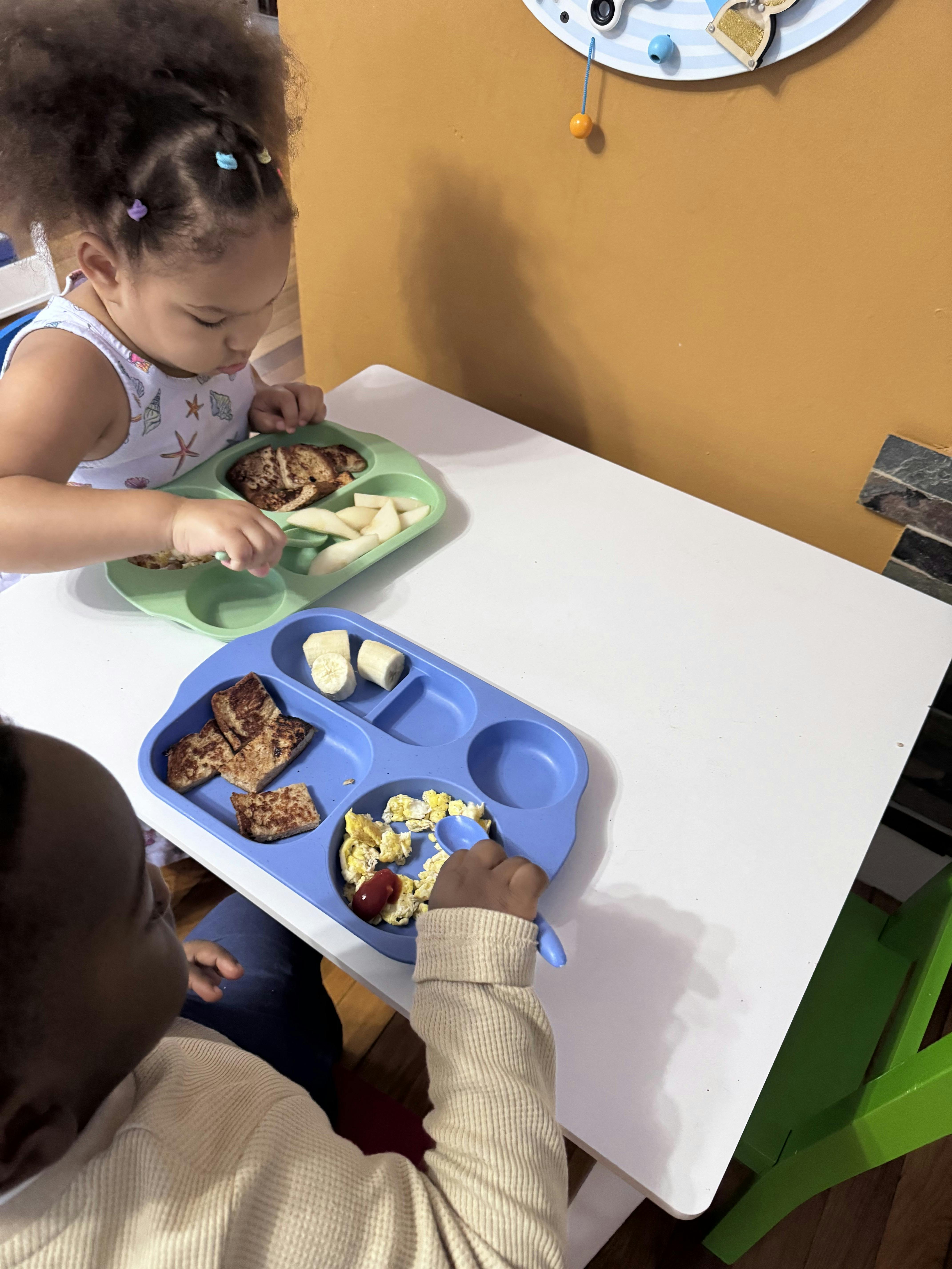 Two children sitting at a small table eating meals from divided trays