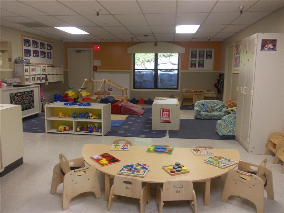 An empty classroom with small wooden tables and chairs, toys, and blue rugs