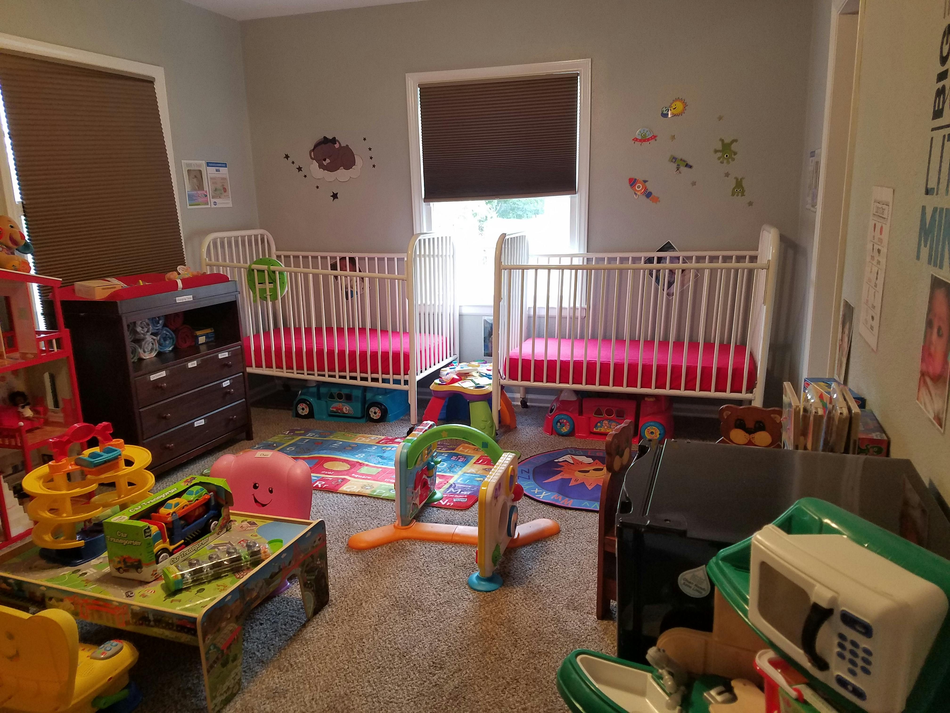 Two cribs with pink bedding in a room with toys and a dresser
