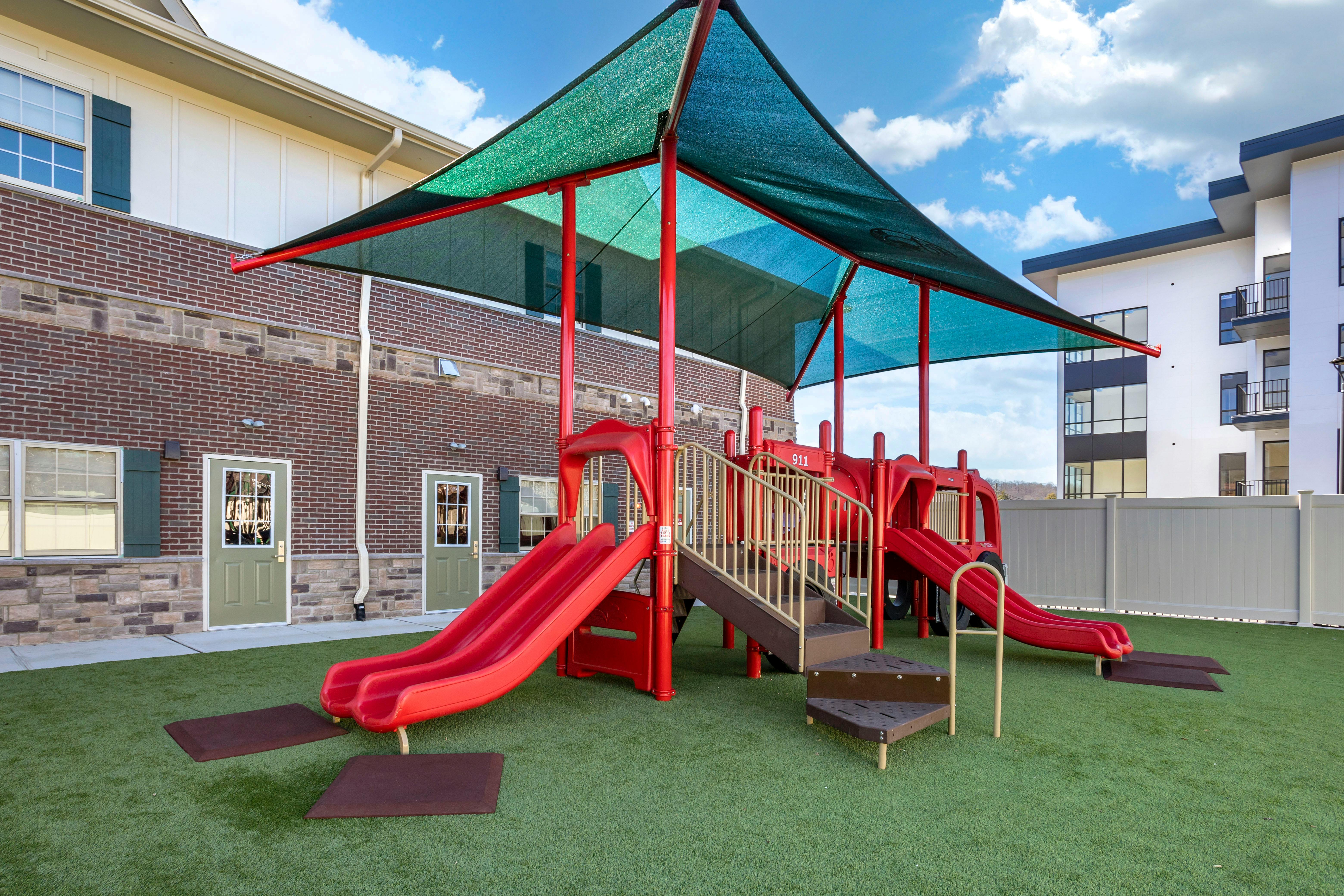 Red playground with slides and steps under a green shade canopy on artificial turf