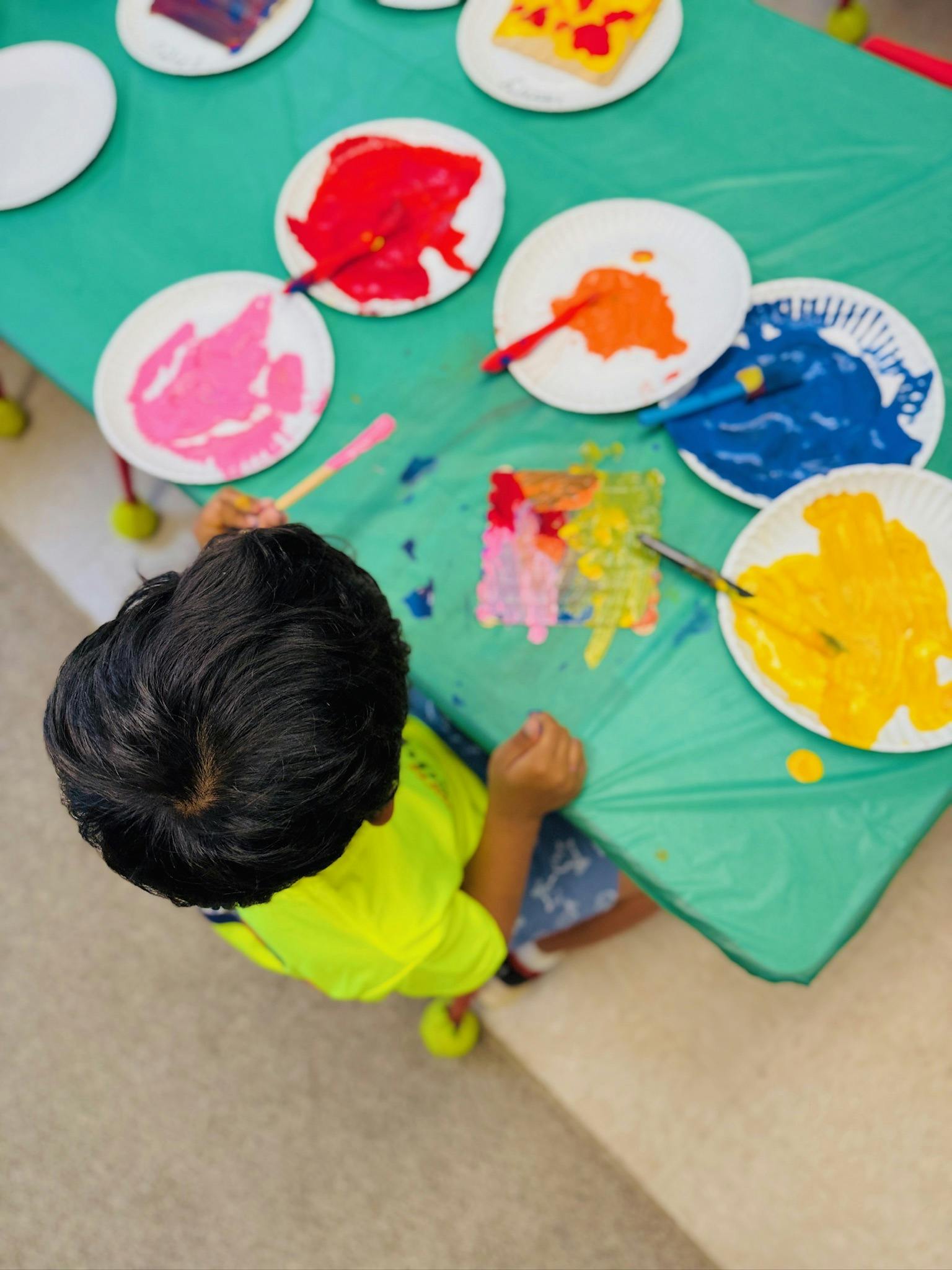 Child painting on paper with various paint colors on a table