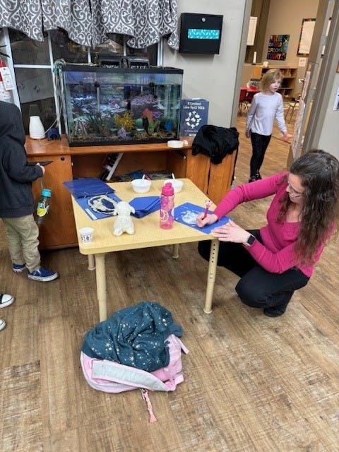 Person kneeling and writing at a small table with a child standing nearby