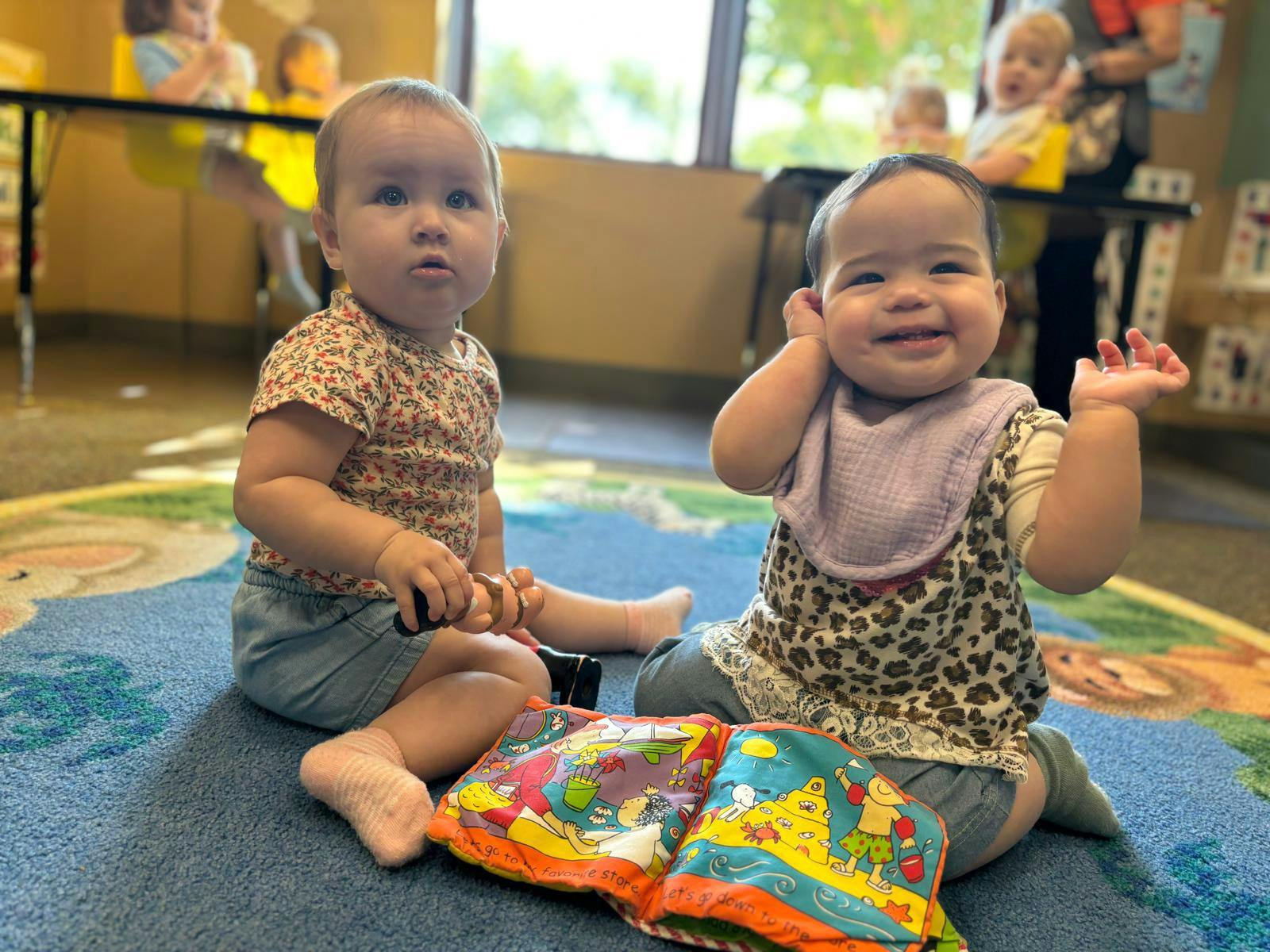 Two babies sitting on a rug with a colorful book