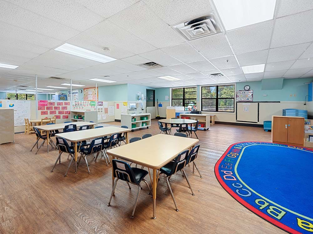 Empty classroom with small tables and chairs, and a blue rug with alphabet letters