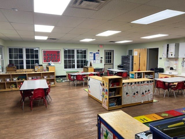 Empty classroom with small tables and chairs, shelves with toys, and bulletin boards