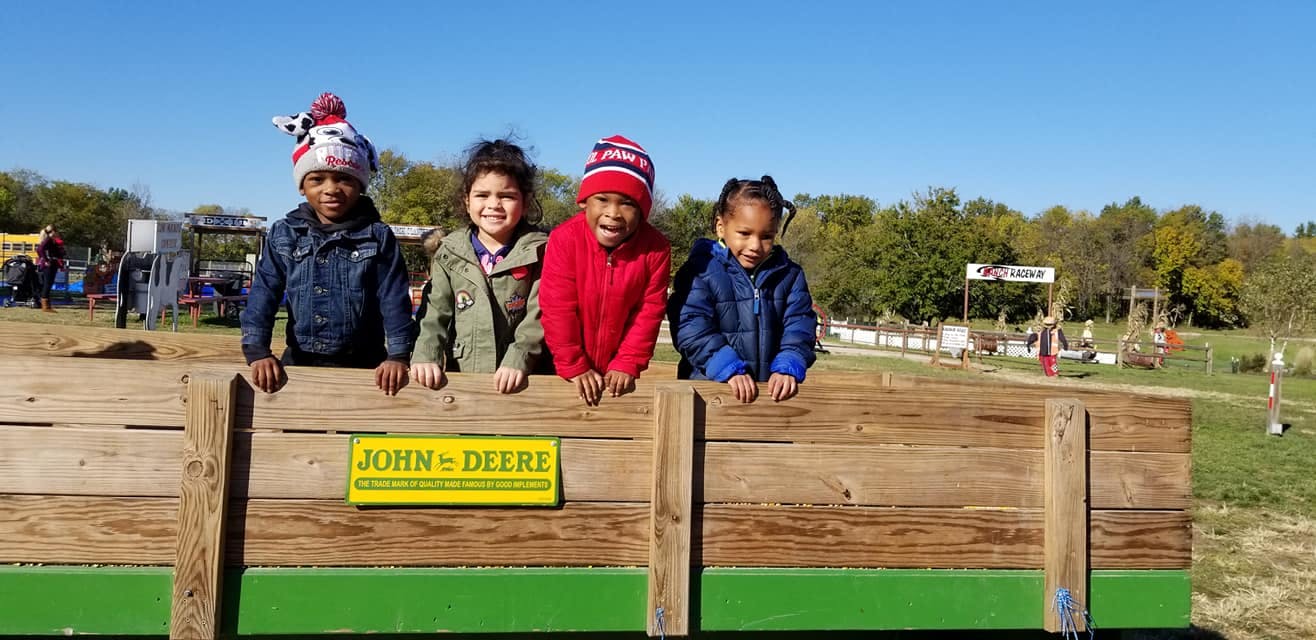 Four children in winter hats looking over a wooden fence with a John Deere sign