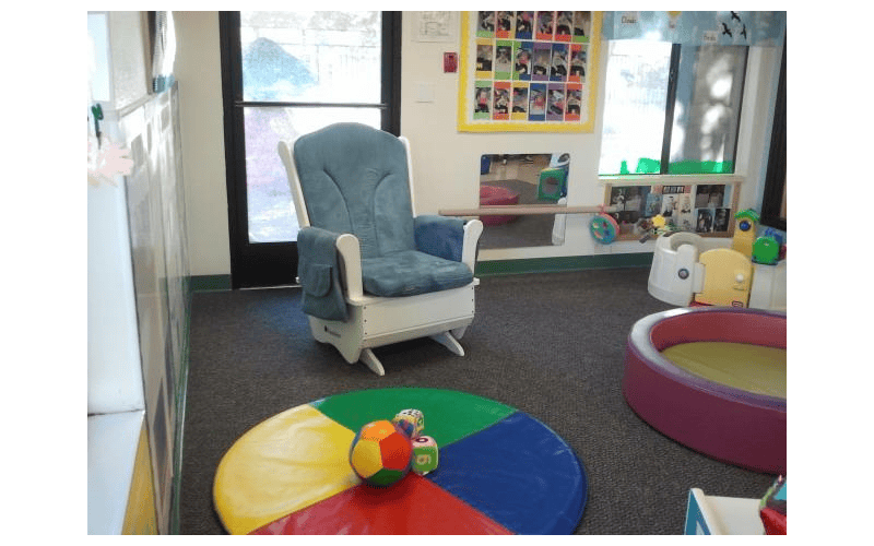 A classroom with a blue glider chair, a colorful mat with a ball, and a pink play pit