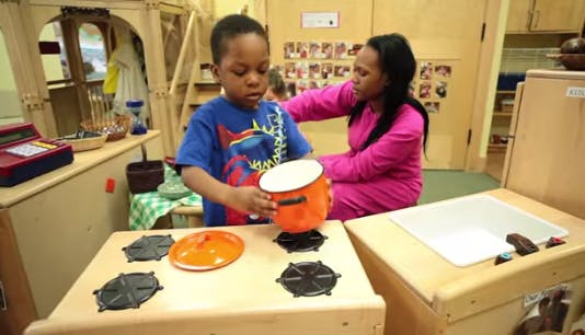 Child playing with toy pot on a play stove with an adult nearby