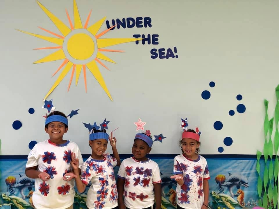 Four children in decorated shirts and headbands in front of a wall with "Under the Sea"