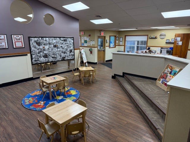 Classroom space with small tables and chairs, a play rug, and a reception desk
