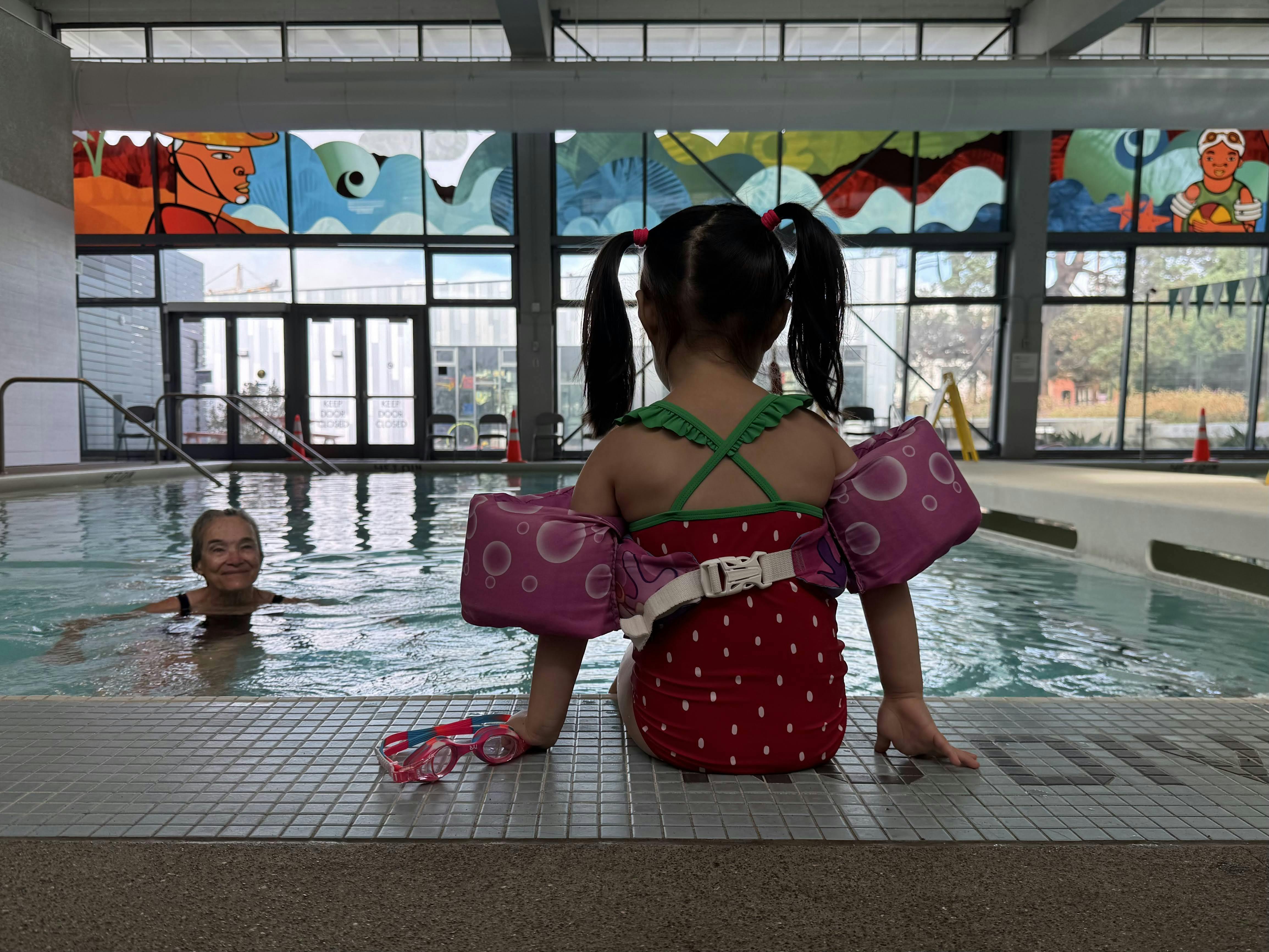 Child in arm floaties sitting at edge of pool, person swimming in background