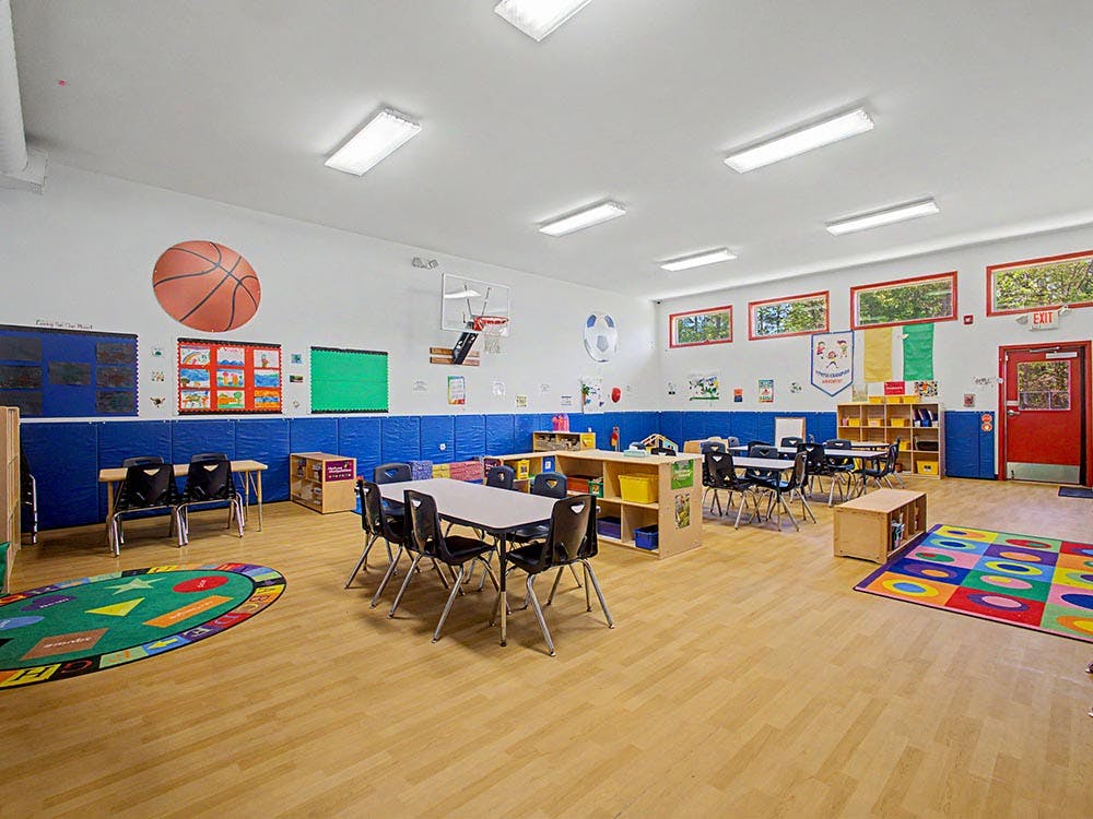 Empty classroom with small tables and chairs, a basketball hoop, and colorful rugs