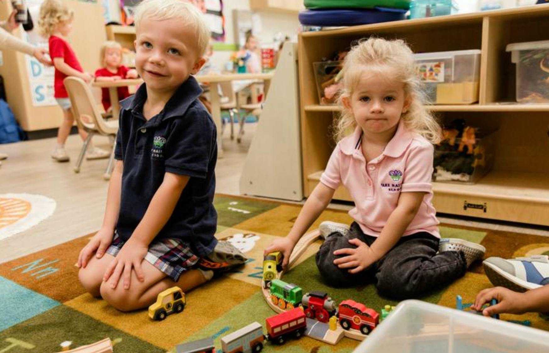 Child kneeling on a rug next to another child playing with a toy train