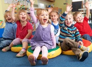 Six children sitting on cushions with hands raised