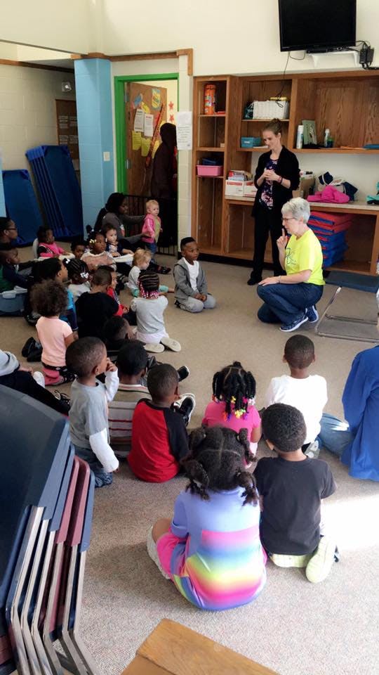 Adults speaking to a group of children sitting on the floor in a classroom