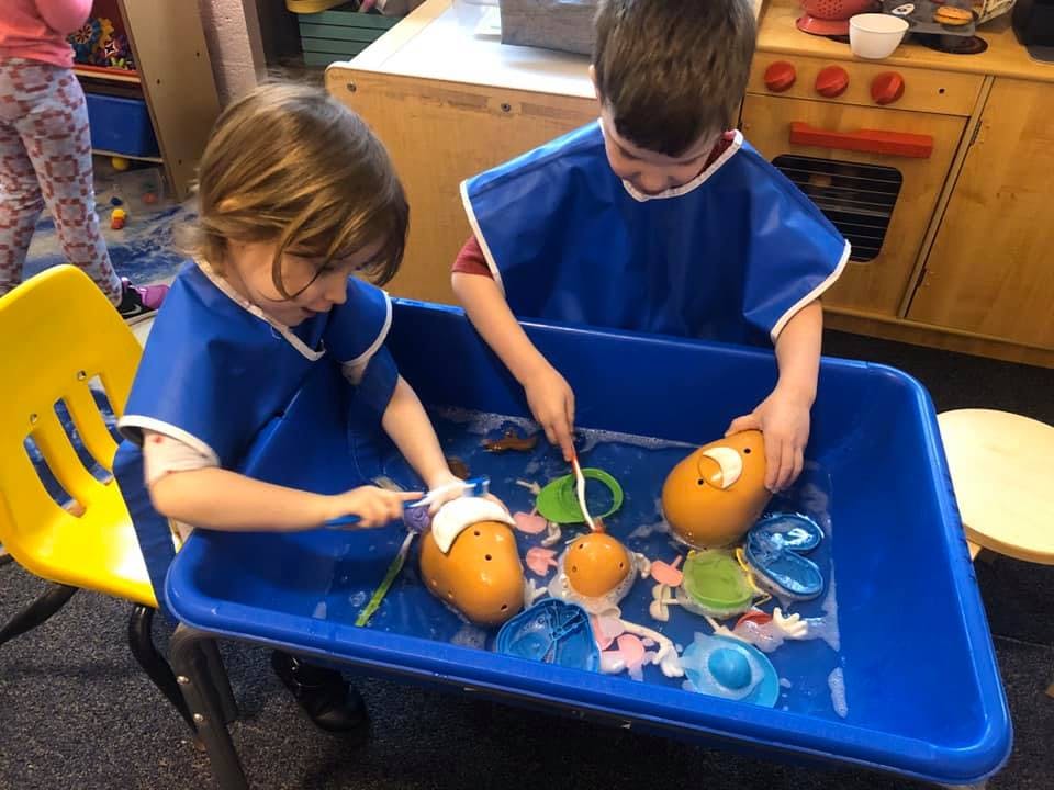 Two children in blue smocks playing with toys in a water table