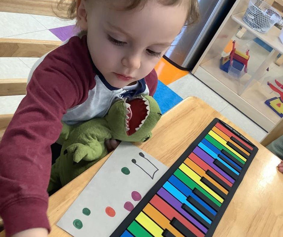 Child with a stuffed dinosaur looking at a colorful toy piano