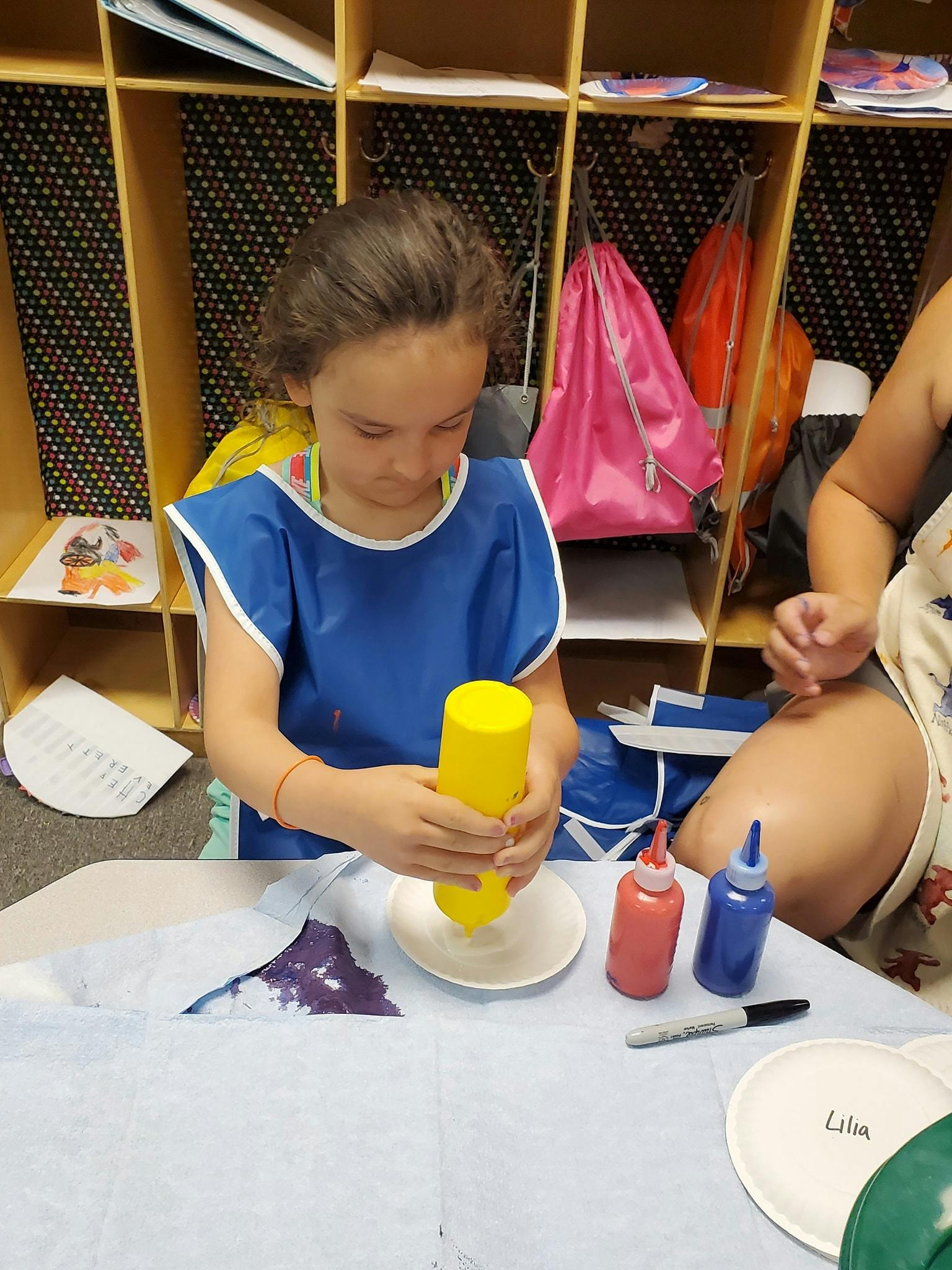Child in a blue smock squeezing a yellow bottle over a plate