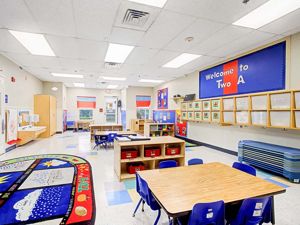 Empty classroom with small tables and chairs, storage shelves, and a welcome sign