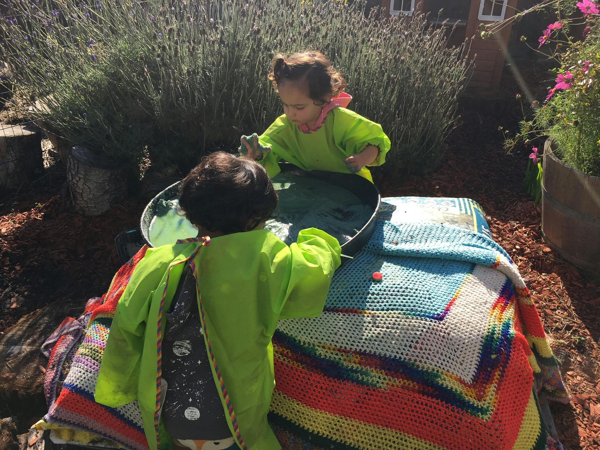 Two children playing with a metal basin on a colorful blanket outdoors